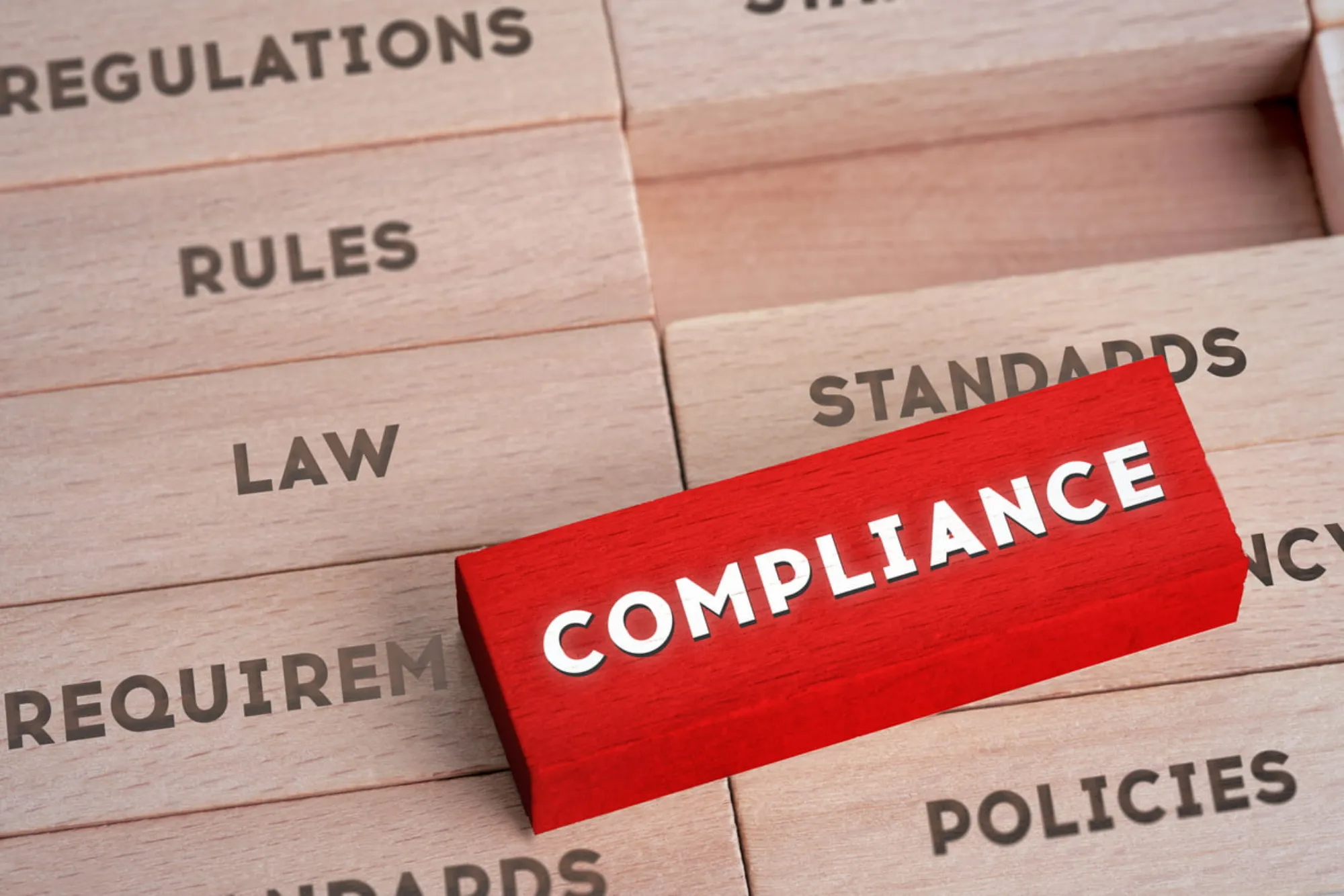 A red wooden block with the text "Compliance" on top of other wooden blocks with text related to rules and regulations.