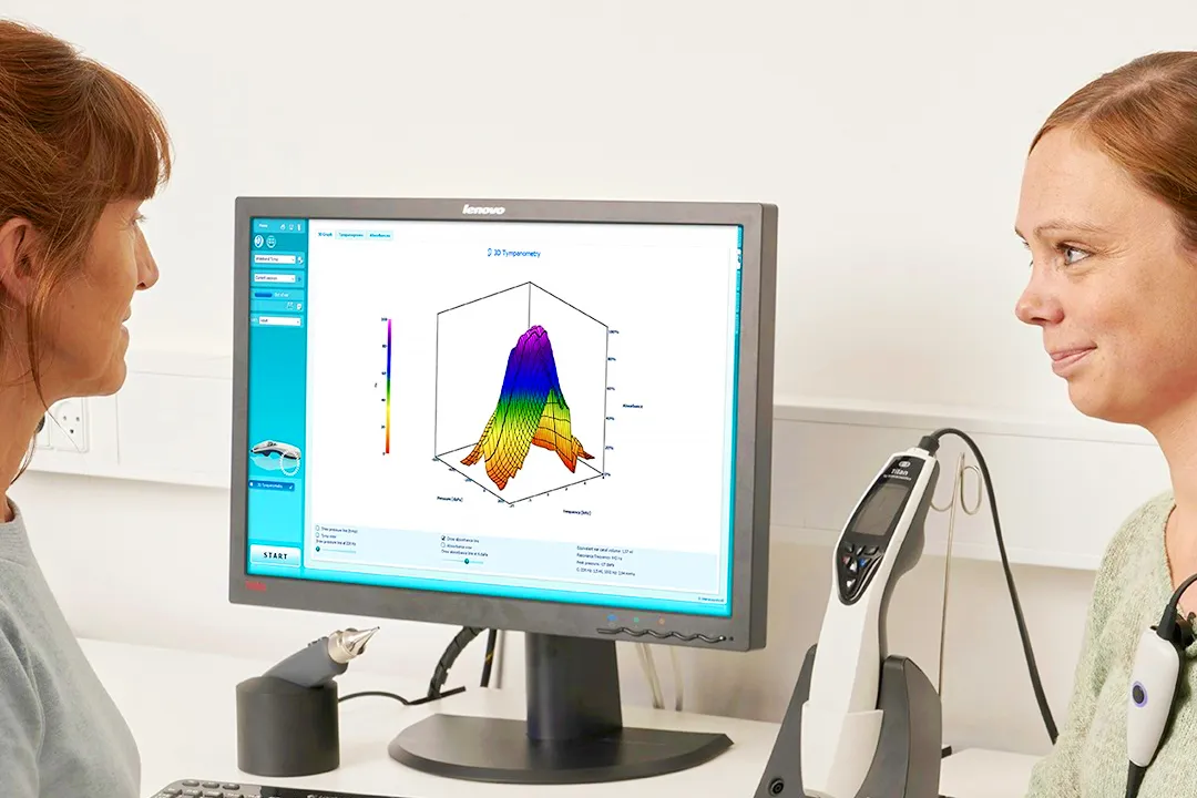 Practitioner explaining a graph on a monitor to a patient with a tympanometer on the desk in the background.